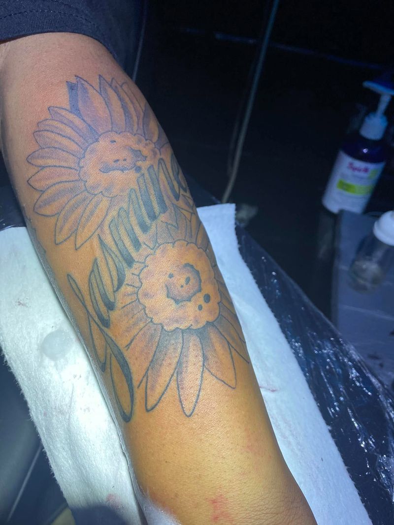 Black and grey tattoo of two sunflowers with flowing script between them on an arm.