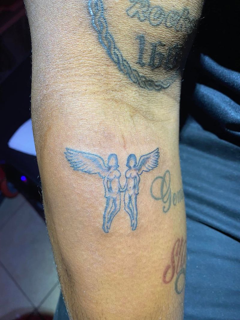 Black and grey tattoo of two winged angel figures standing side by side on a forearm.